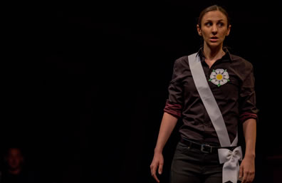 York, in dark purple shirt, black pants, a white rose on her shirt and a white sash with a bow at her belt, stands to one side of the photo, looking off to the side warily.